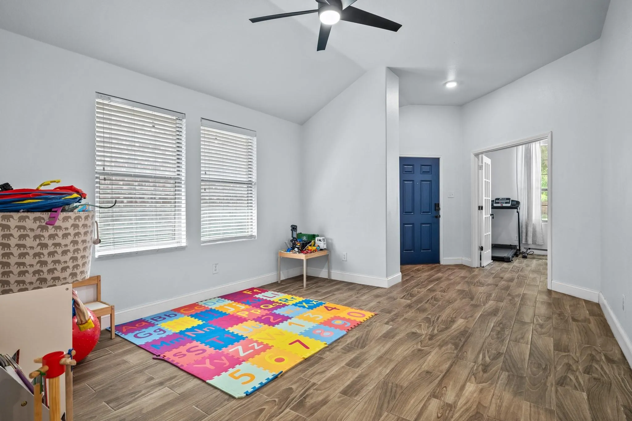 Vaulted ceiling with a ceiling fan, two windows with blinds, wood-finish flooring, and a paneled door