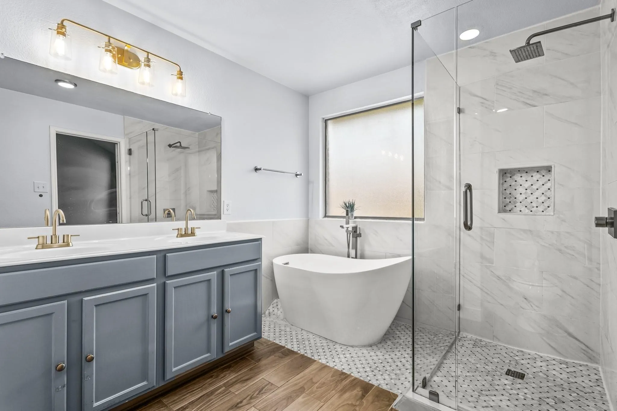 Dual vanity with gold-tone fixtures, wood-finish flooring, freestanding soaking tub, glass-enclosed shower with rain showerhead, and a tiled shower niche