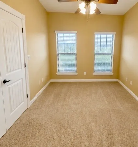 Neutral tone wall paint, carpeted flooring, two windows with blinds, a white paneled door with dark hardware, and a ceiling fan with integrated lighting