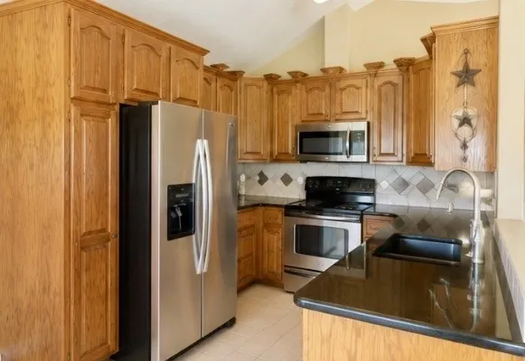 Kitchen featuring wood cabinetry, stainless steel appliances, dark granite countertops, a tile backsplash, and a built-in sink with a gooseneck faucet