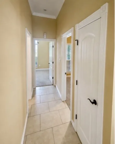 Hallway featuring neutral wall paint, tile flooring, white crown molding, and white doors with dark hardware