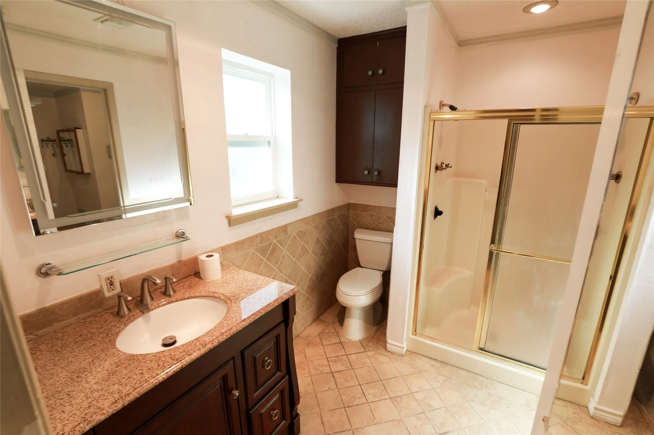 Vanity with stone countertop and undermount sink, built-in storage cabinetry, tiled shower with gold-tone frame, and floor tiling