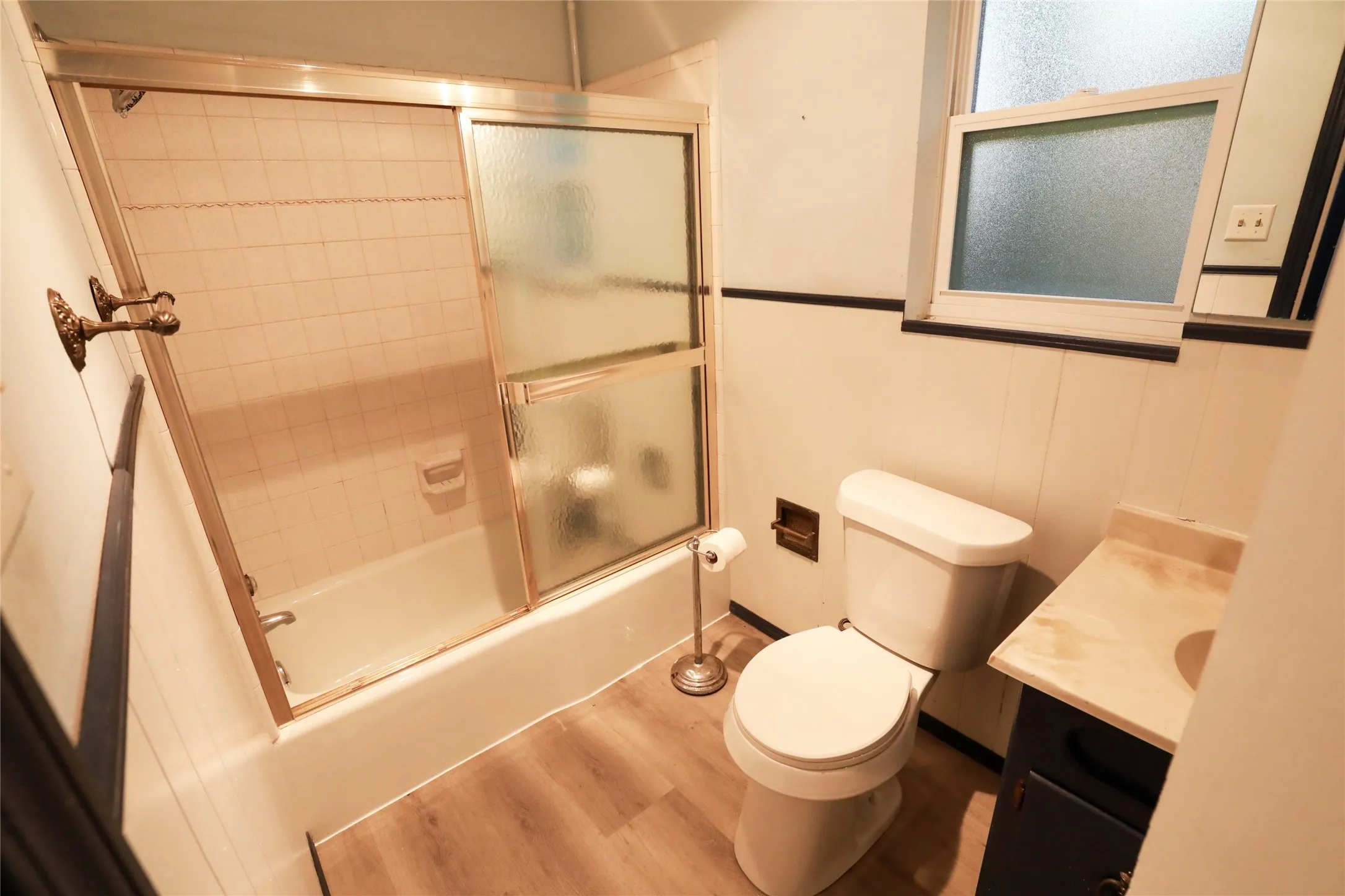 Bathtub with sliding glass enclosure, white tile surround, wood-finish flooring, and a window with frosted glass