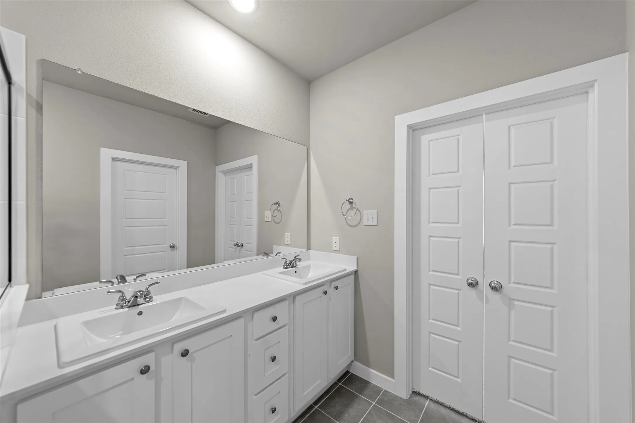 Dual undermount sinks with chrome fixtures, expansive vanity with white cabinetry, large wall-mounted mirror, and gray floor tile