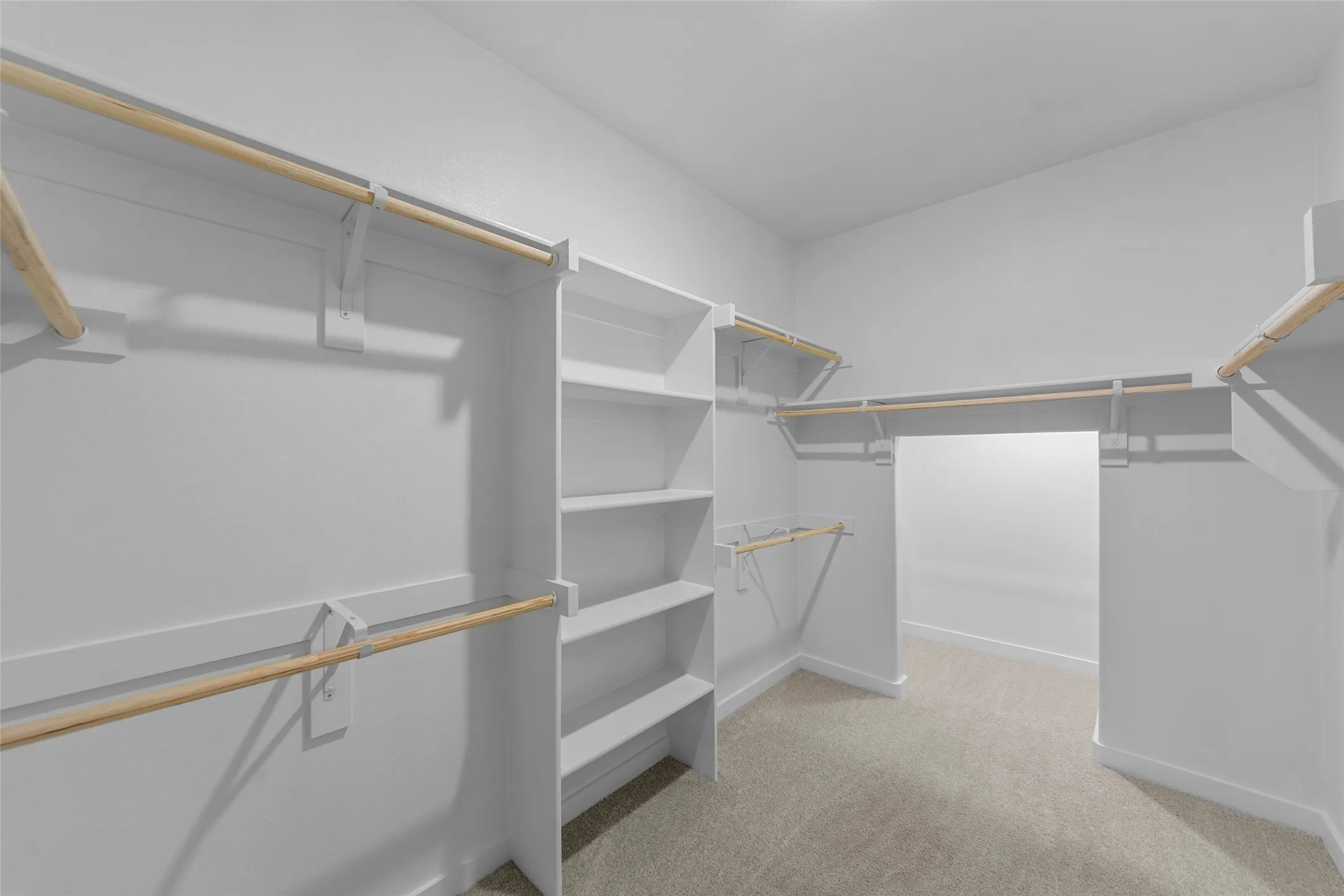 Spacious walk-in closet featuring white built-in shelving, multiple hanging rods, and light gray carpeting
