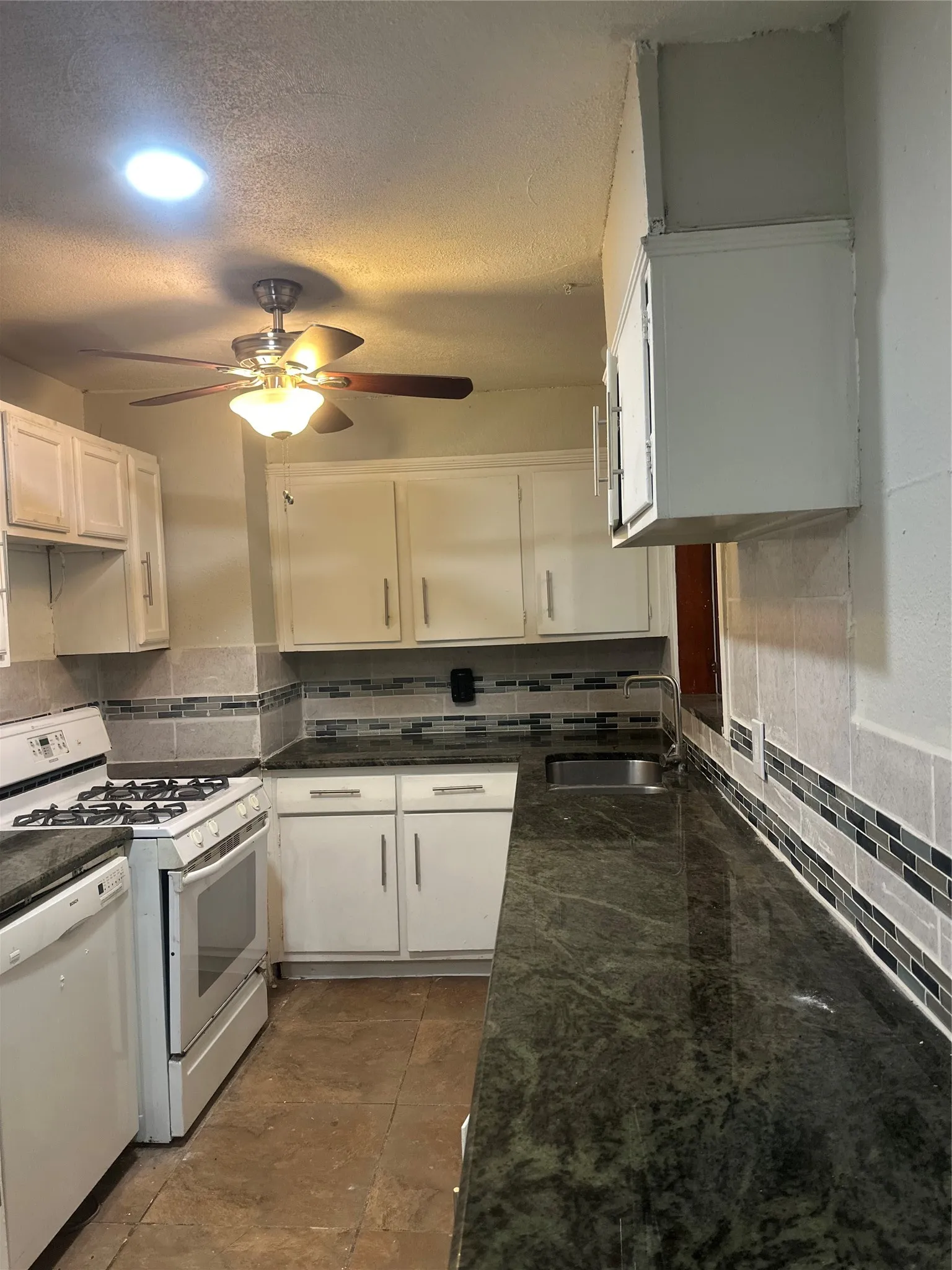White cabinetry, dark stone countertops, tile backsplash, built-in range, and a ceiling fan with integrated lighting
