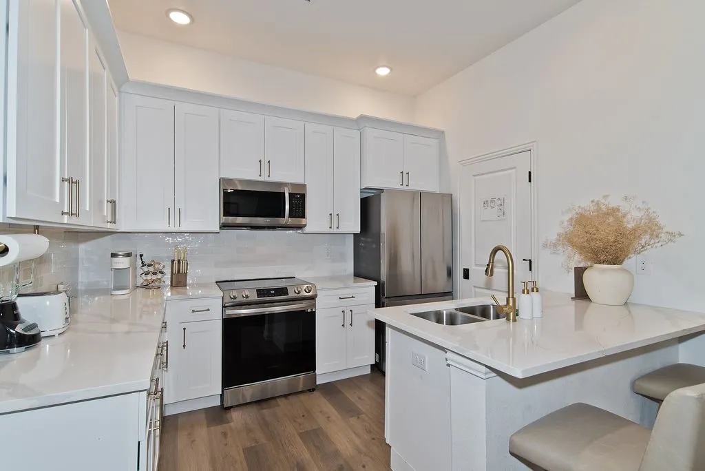 Modern kitchen featuring white shaker-style cabinetry, stainless steel appliances including a gas range and built-in microwave, quartz countertops with waterfall edge at the island, a bronze pull-down faucet, subway tile backsplash, and wood-finish flooring throughout