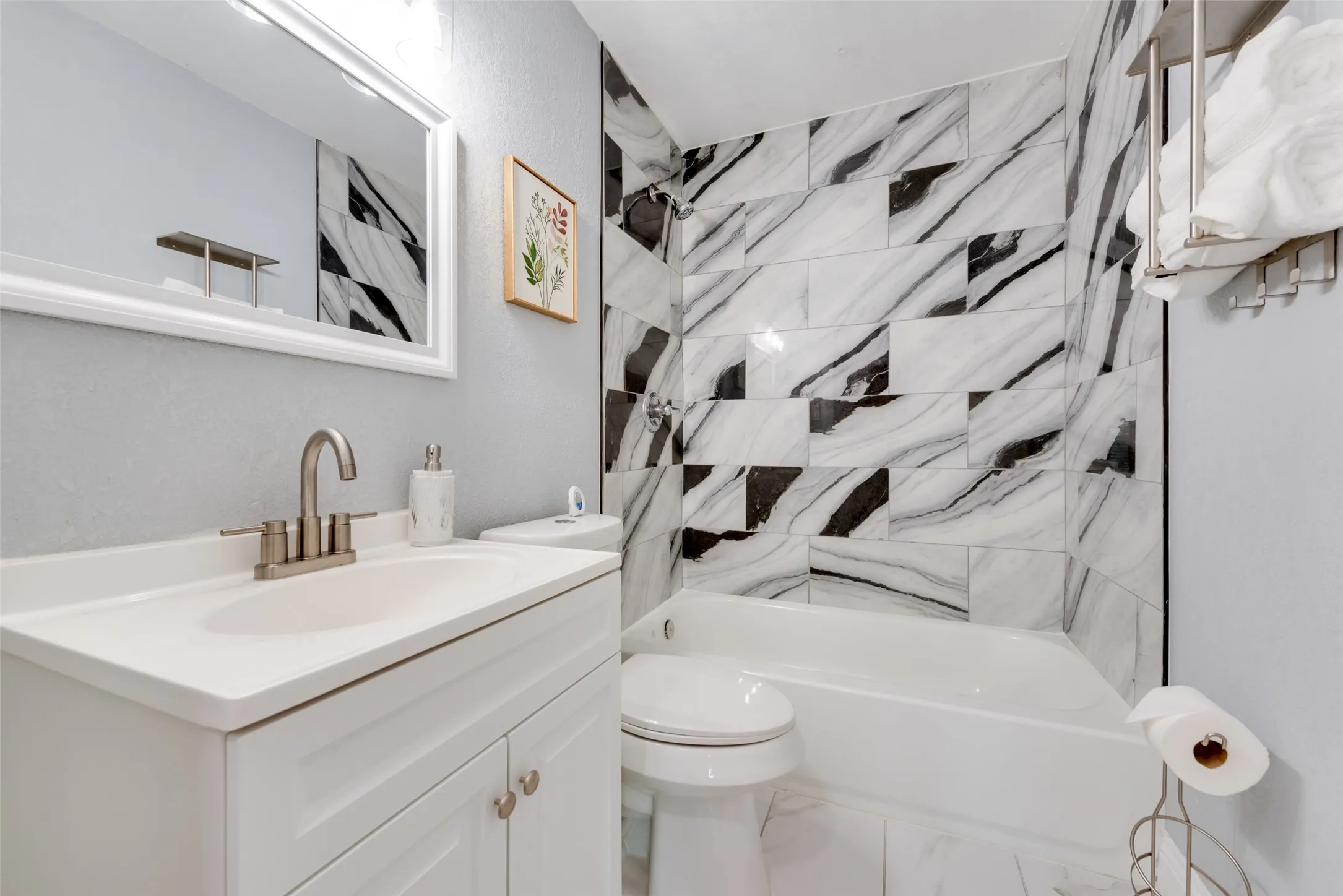 White vanity with integrated sink, brushed nickel faucet, framed mirror, and a tiled shower/tub combination with contrasting black and white patterns