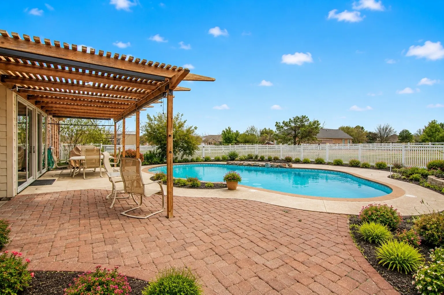 Expansive brick paver patio with a wooden pergola, leading to a curved swimming pool