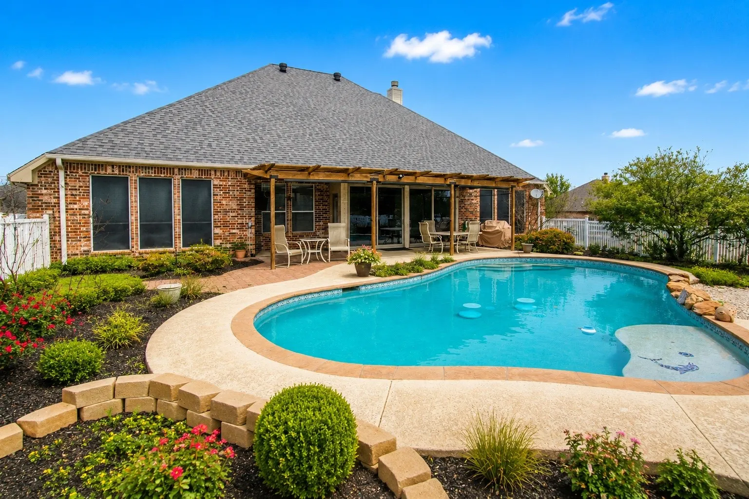 Brick exterior with multiple windows, covered patio, a shaped pool with integrated steps, and landscaped garden beds
