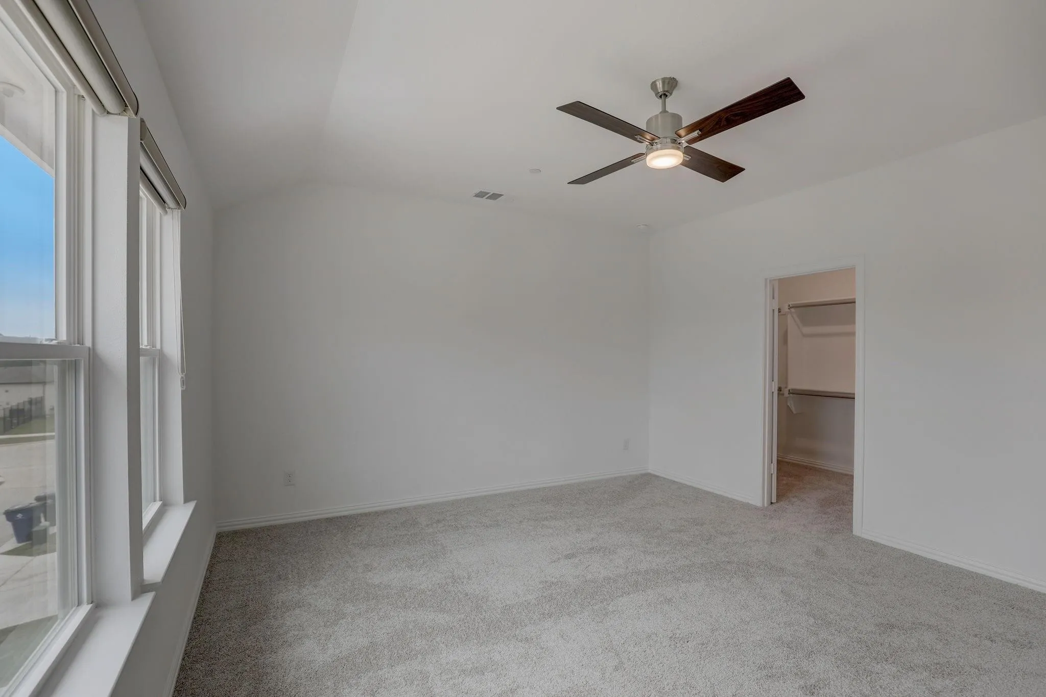 Vaulted ceiling, ceiling fan with light, gray carpet, multiple windows with blinds, and a walk-in closet with built-in shelving