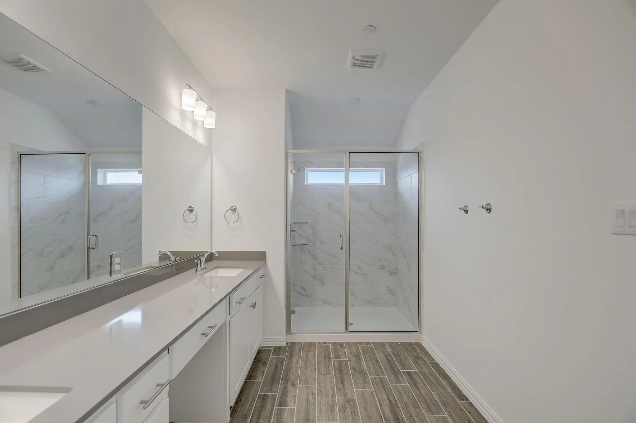 Dual vanity with integrated sinks, expansive mirror, modern light fixture, glass-enclosed shower, and wood-finish tile flooring