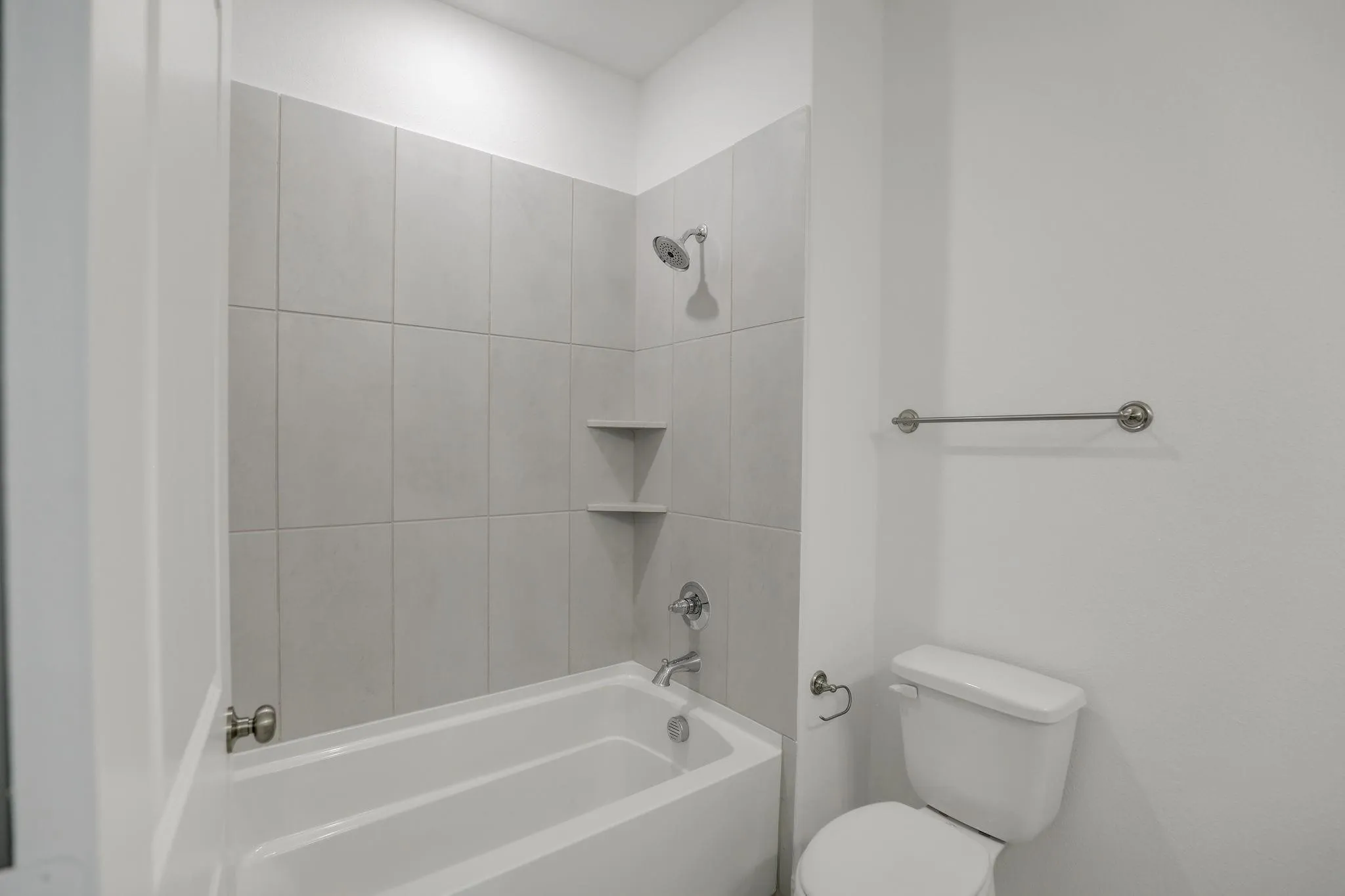 White bathtub with chrome shower fixtures, tiled shower surround, and integrated corner shelving
