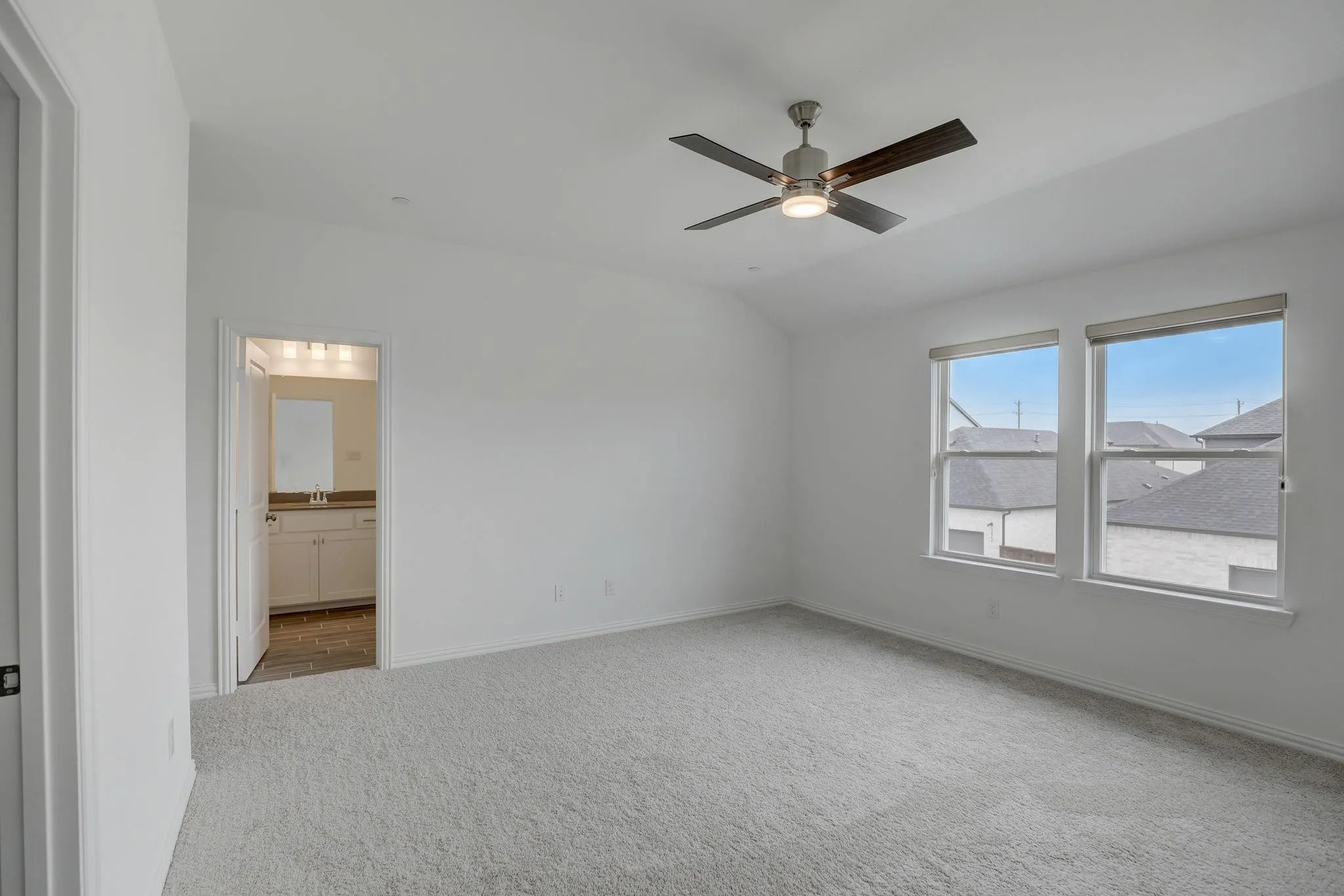 Vaulted ceiling with a ceiling fan, two windows, carpet flooring, and an open doorway to a bathroom with a vanity and mirror