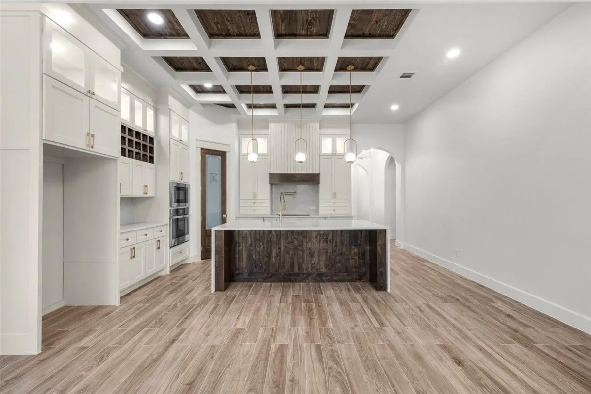 Coffered ceiling with wood-finish panels, white shaker cabinetry with gold-tone hardware, wood-finish flooring, central island with light-toned countertop, and built-in double ovens