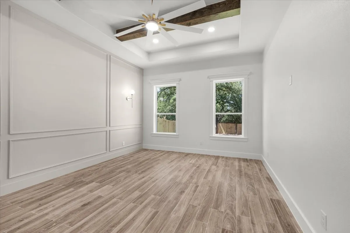 Tray ceiling with exposed wood beams, ceiling fan, wood-finish flooring, two windows, and decorative wall molding
