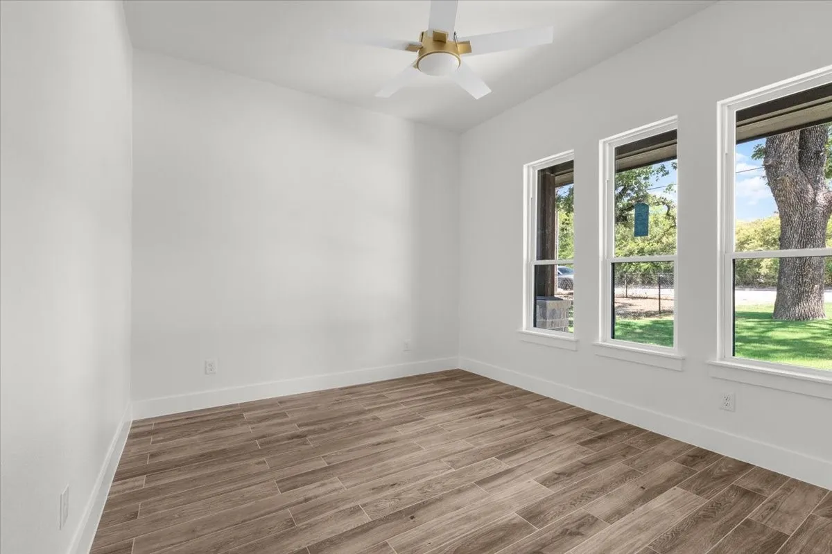 Wood-finish flooring, white walls, three large windows, white window frames, and a ceiling fan with a gold-tone fixture