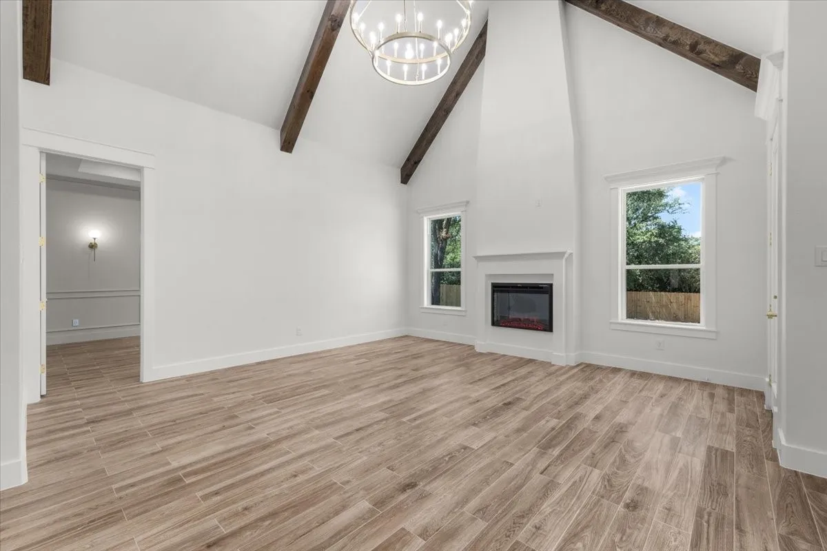 Vaulted ceiling with exposed wood beams, wood-finish flooring, two large windows, built-in fireplace, and a contemporary chandelier