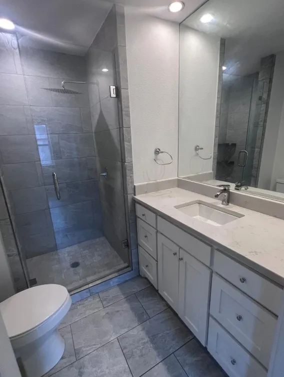 Glass-enclosed shower with a rainfall showerhead, white vanity with stone countertop, integrated sink, and recessed lighting
