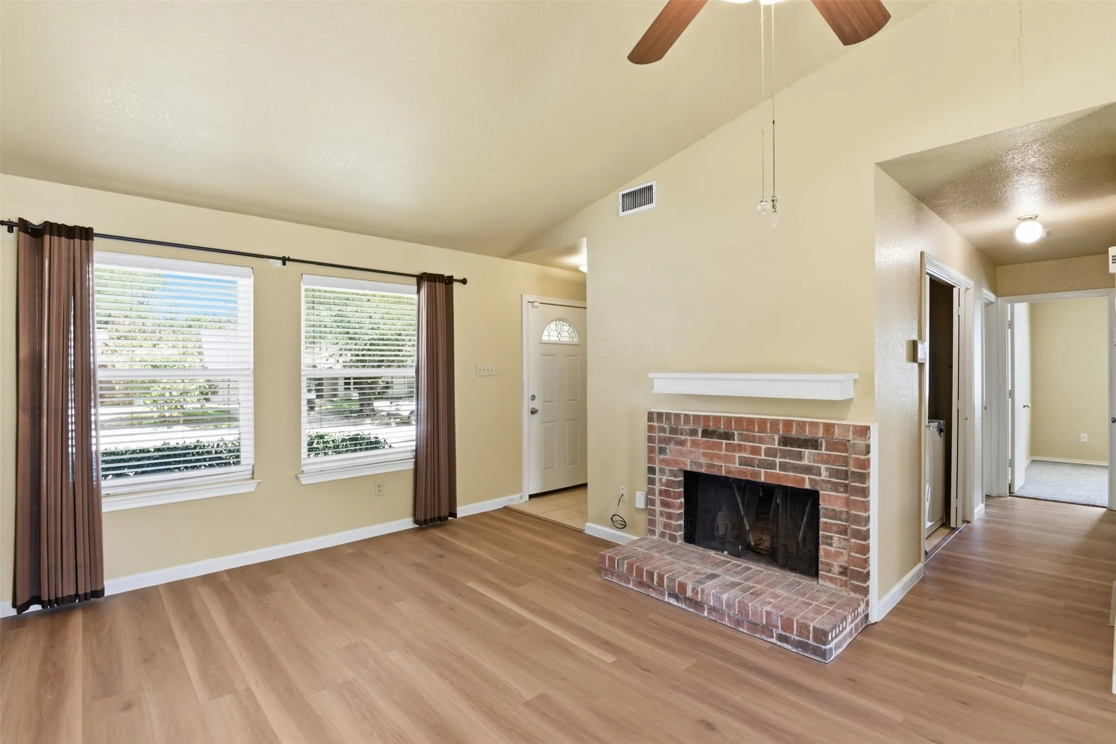 Wood-finish flooring throughout, brick fireplace with a white mantel, dual windows with blinds, a vaulted ceiling, and an overhead ceiling fan