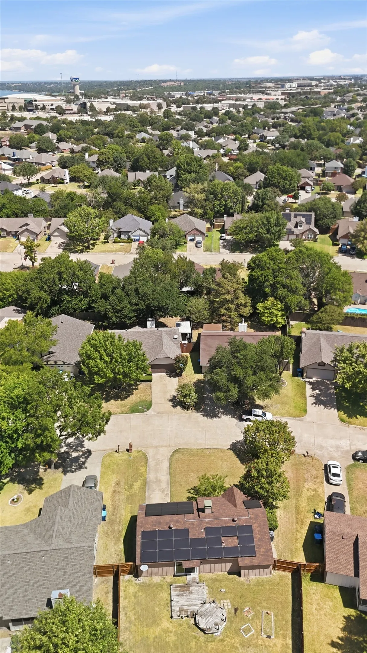 Residential neighborhood with mature trees, a subject property featuring a brown roof, solar panels, and a fenced backyard