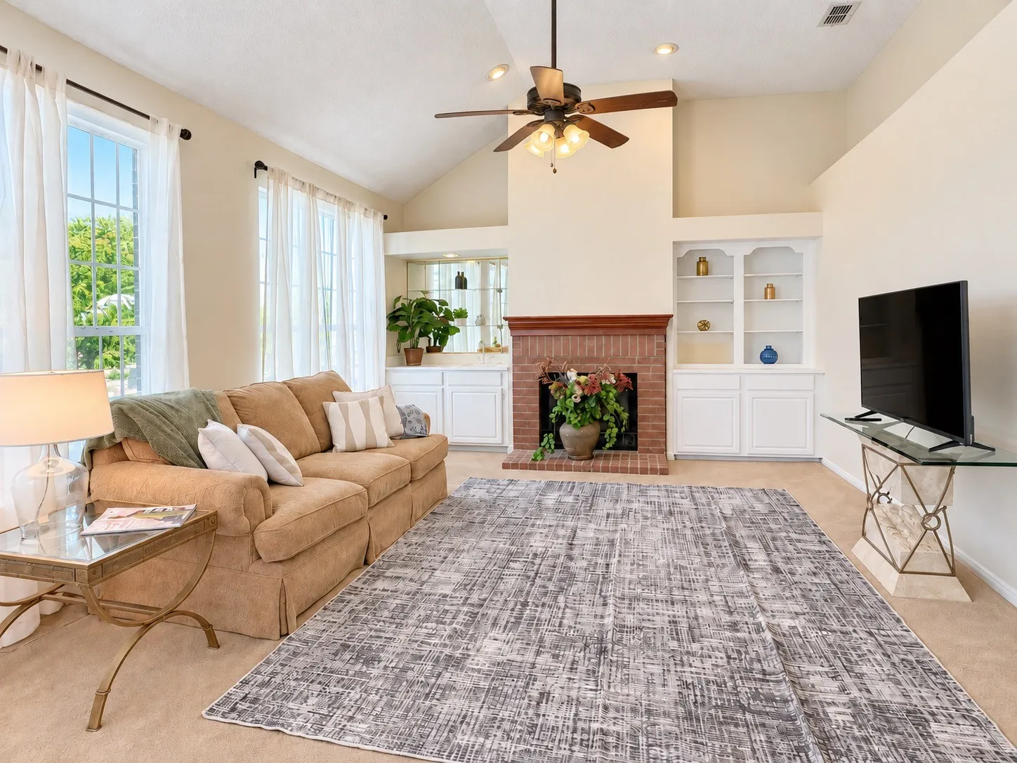 Vaulted ceiling with a ceiling fan, brick fireplace, built-in shelving, and large windows