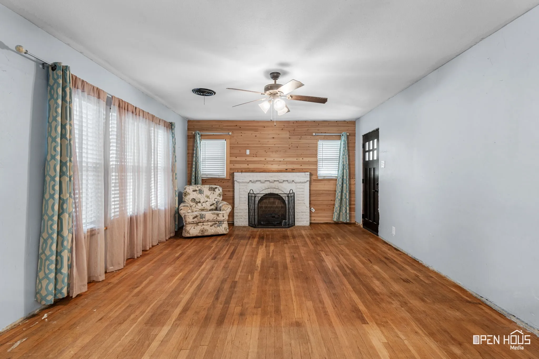 Hardwood flooring, white brick fireplace with hearth, wood-paneled accent wall, ceiling fan, and multiple windows