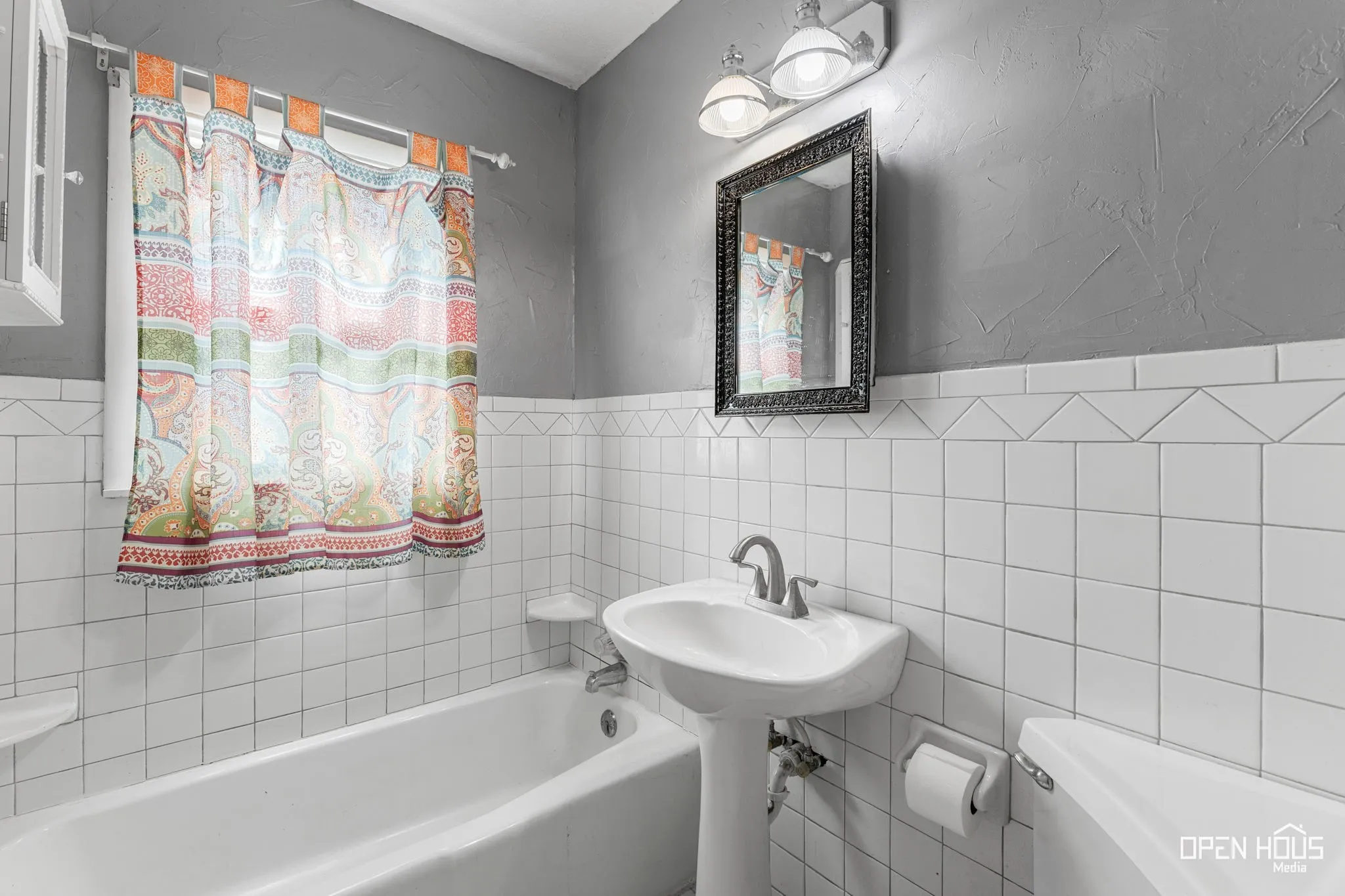 Pedestal sink, wall-mounted light fixture, decorative framed mirror, white tile wainscoting with patterned accent, and a built-in bathtub