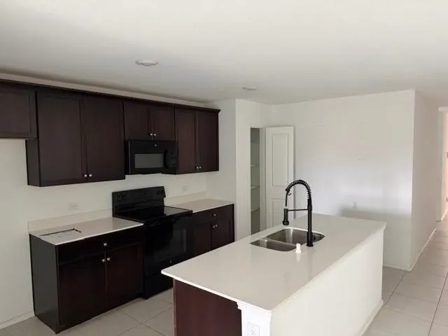 Kitchen featuring dark wood-finish cabinetry, white countertops, built-in microwave, black electric range, and a kitchen island with a double basin sink and matte black faucet