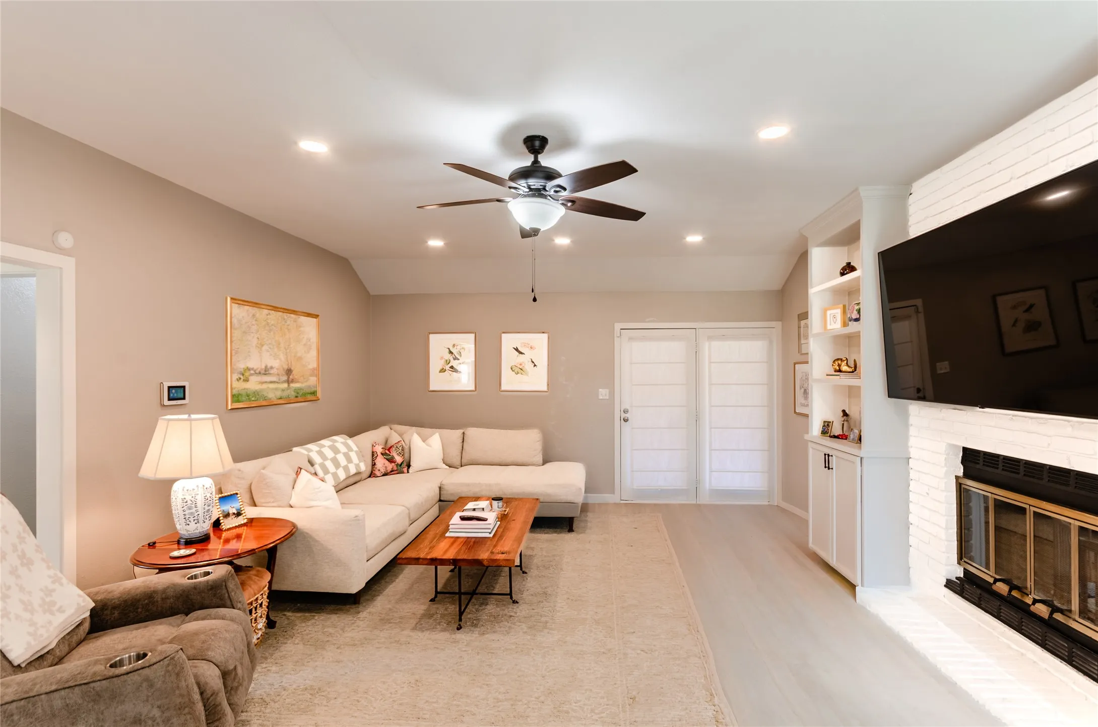 Living area featuring a ceiling fan with light, recessed lighting, a fireplace with a white brick surround, built-in shelving, and wood-finish flooring