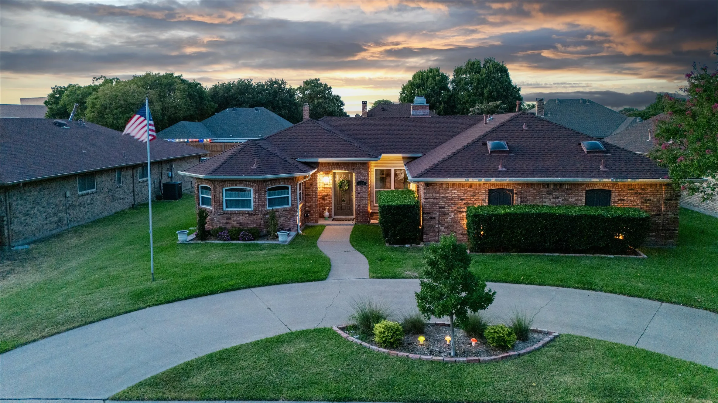 Brick Colonial-style residence featuring a welcoming front entrance with illuminated architectural lighting, manicured lawn with mature landscaping, circular concrete driveway, and an American flag display