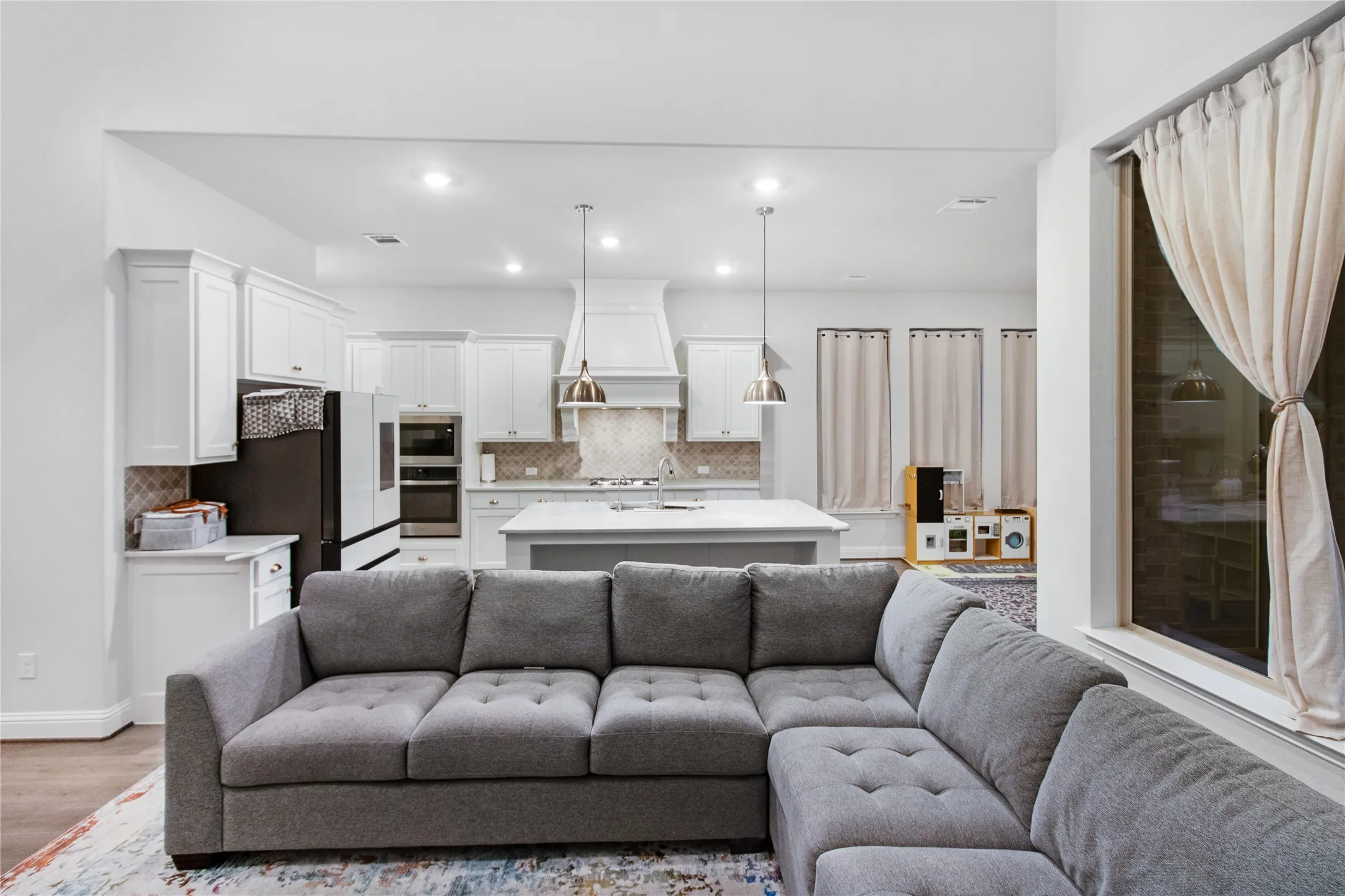 Open-concept living and kitchen featuring a gray upholstered sectional sofa, pendant lighting above the kitchen island, stainless steel appliances, white cabinetry, and recessed ceiling lights throughout the space