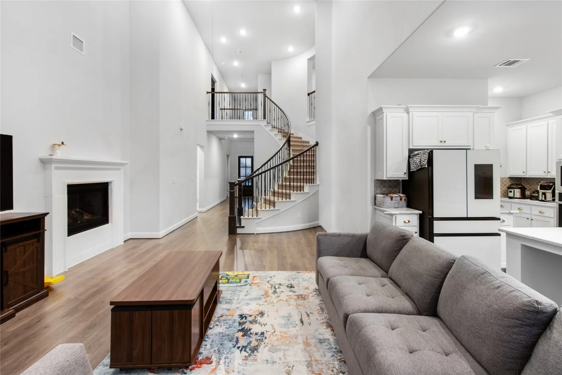 Open-concept living space featuring soaring ceilings with recessed lighting, a curved staircase with metal railings and wood treads, hardwood flooring throughout, a gas fireplace with white surround, and a modern kitchen with white cabinetry and stainless steel appliances