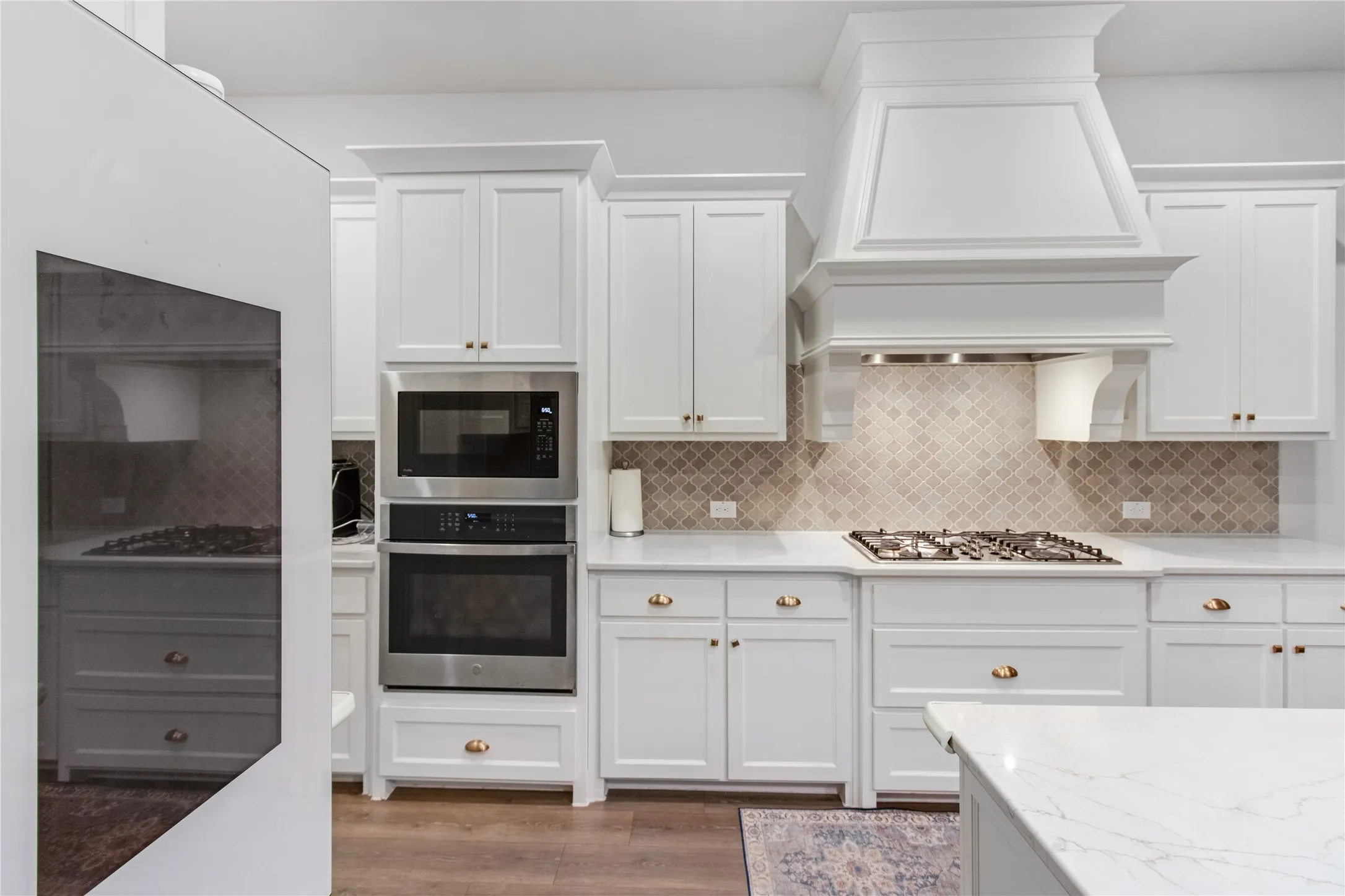 White cabinetry with brass hardware, stainless steel built-in appliances, a gas cooktop, patterned tile backsplash, and light-toned countertops