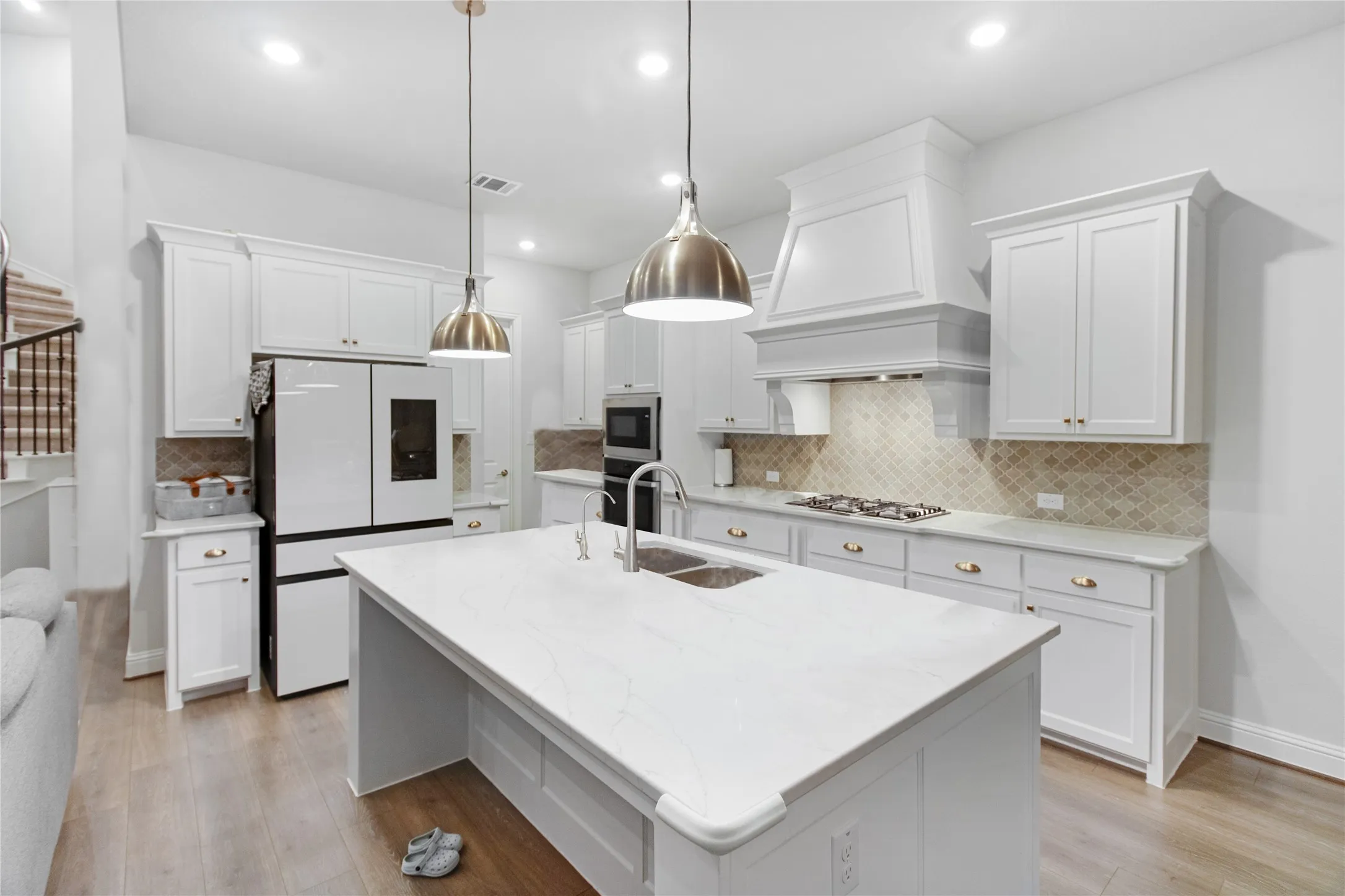 White cabinetry with gold-tone hardware, wood-finish flooring, recessed lighting, and a kitchen island with a double basin sink