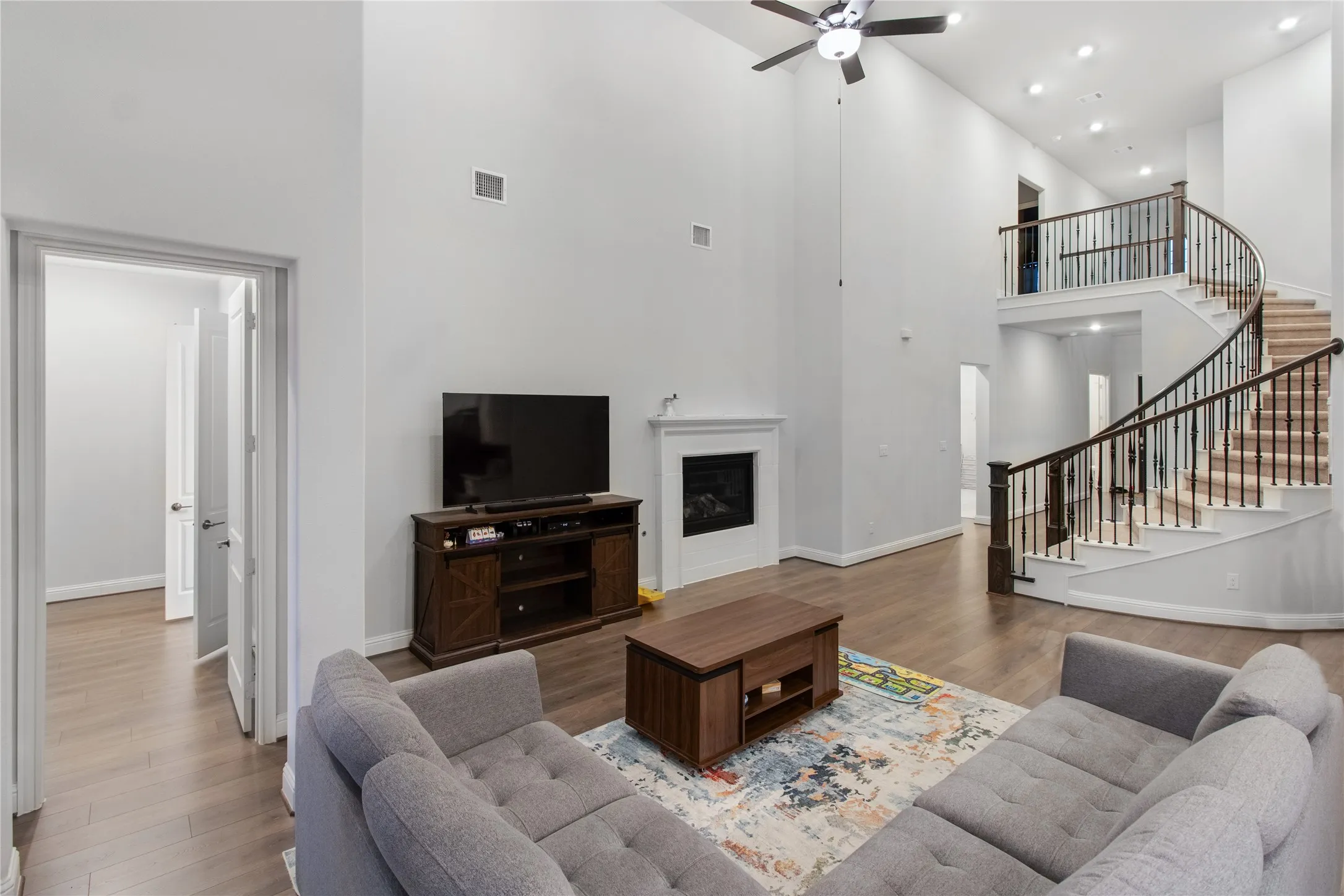 Spacious open-concept living area featuring soaring ceilings with recessed lighting and a ceiling fan, hardwood flooring throughout, and a curved staircase with black metal railings