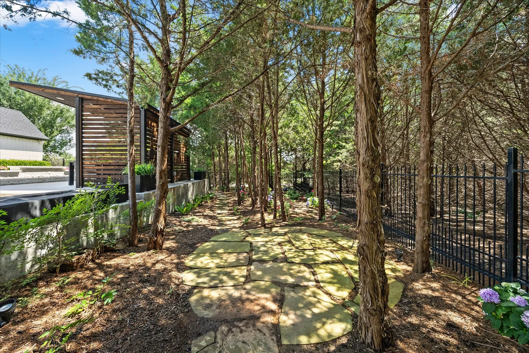 Stone slab pathway with surrounding trees and foliage, featuring a black metal fence and a building with horizontal wood slat siding