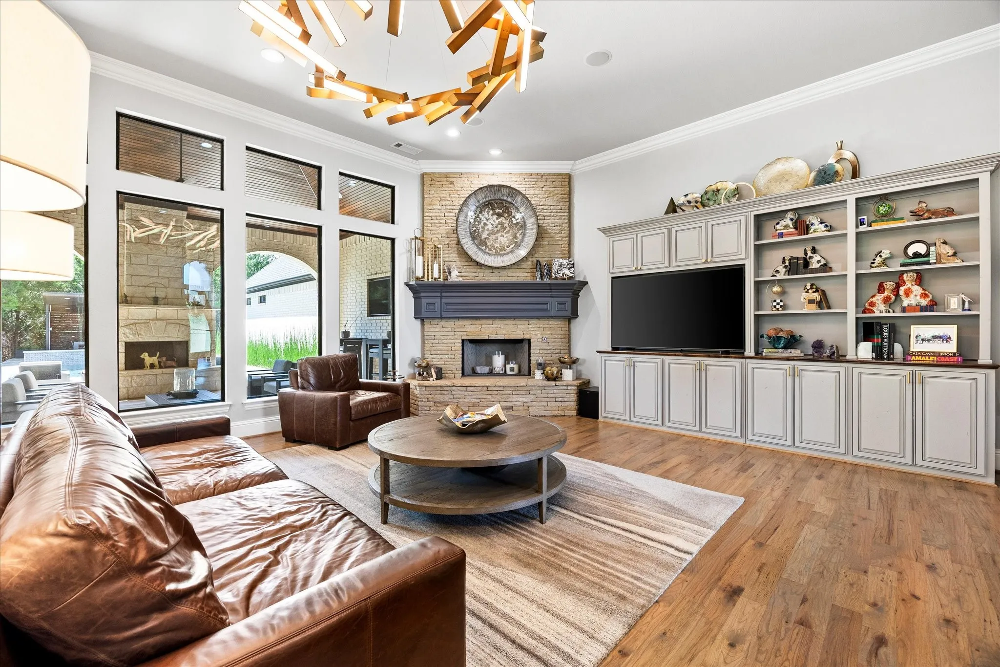 Hardwood flooring, custom built-in media cabinetry, stone-clad fireplace with mantel, crown molding, and expansive windows