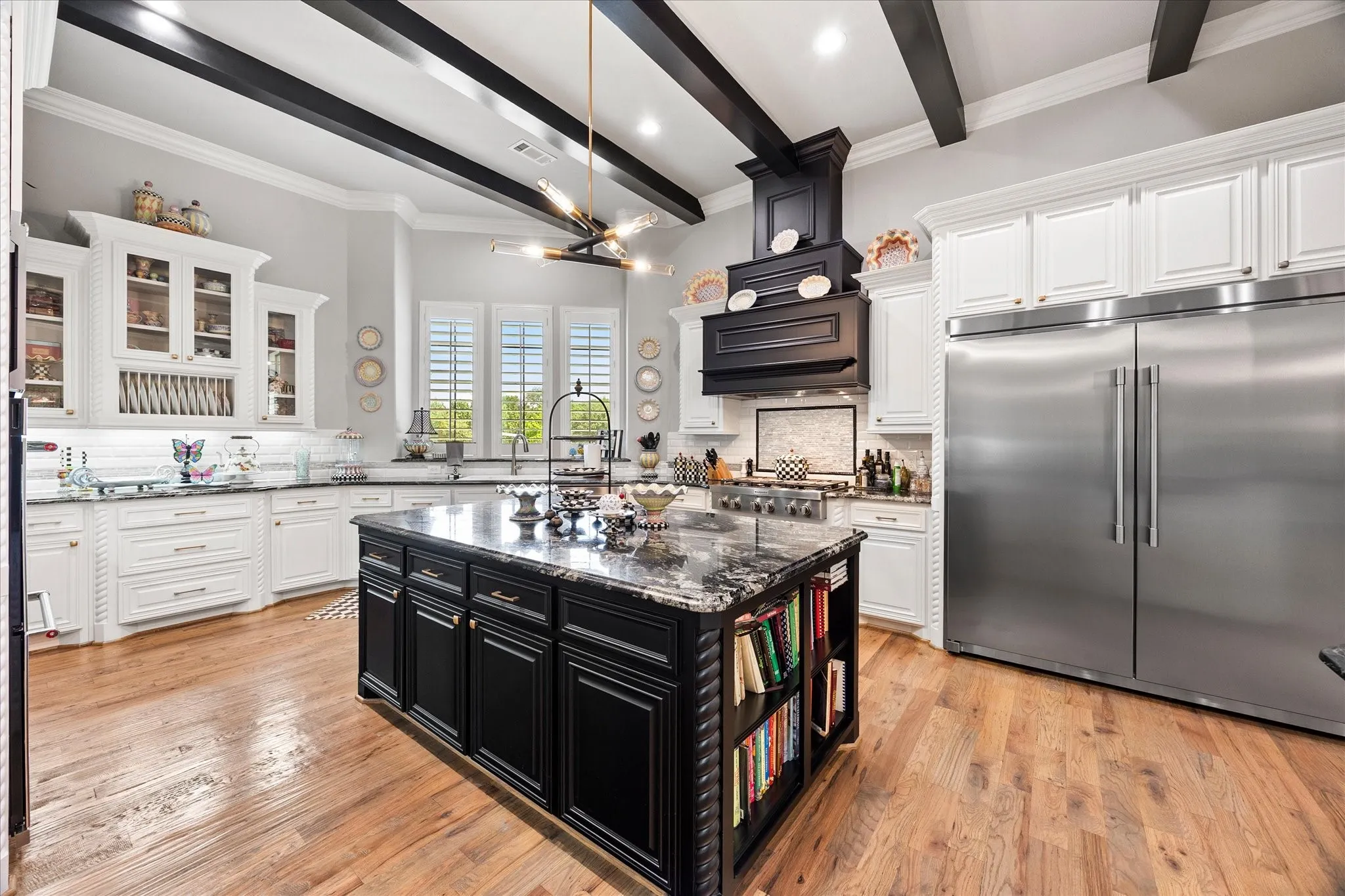 Gourmet kitchen featuring a large central island with storage, wood-finish flooring, white cabinetry, stainless steel appliances, and exposed ceiling beams