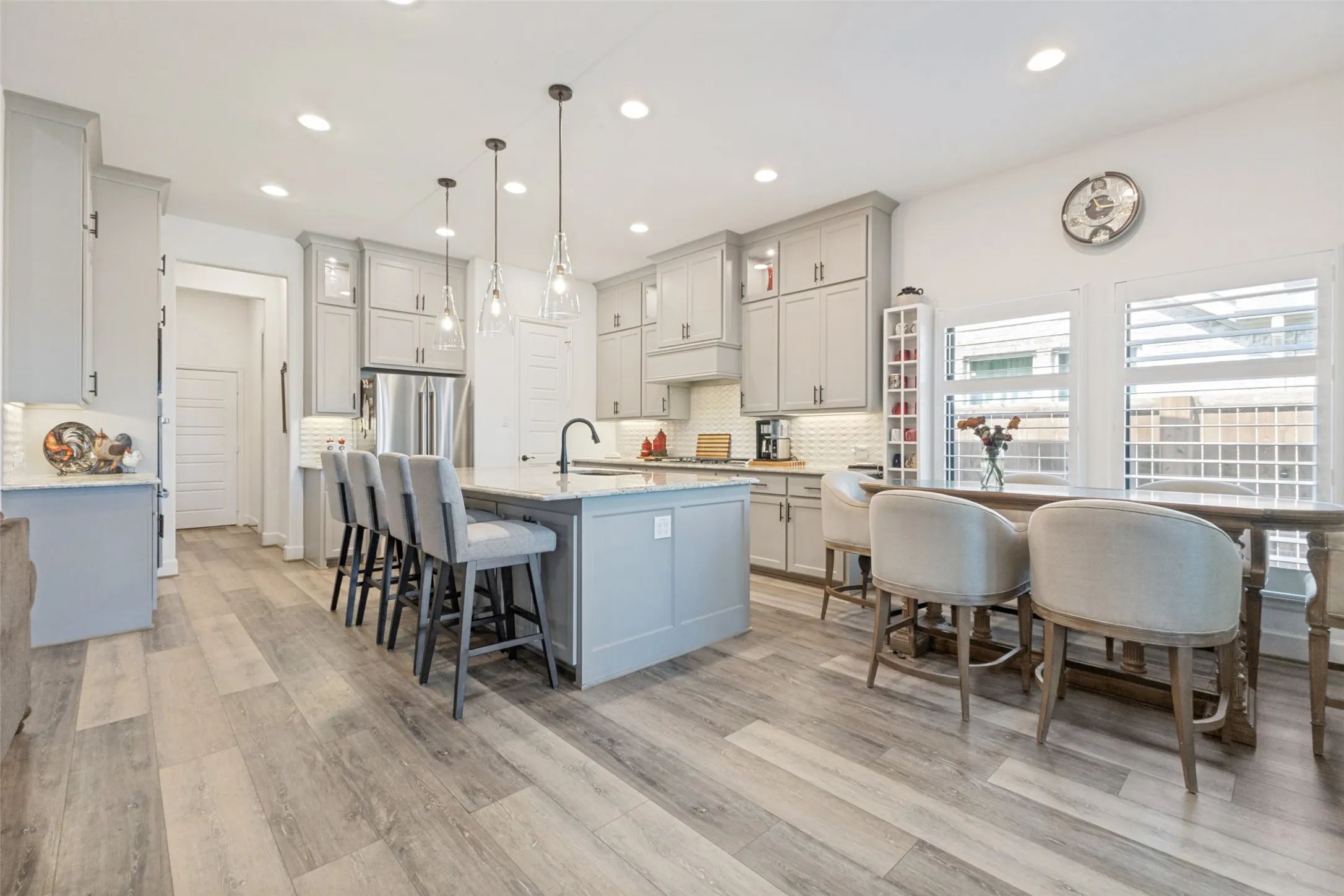 Expansive kitchen featuring light gray cabinetry, stainless steel appliances, a large island with pendant lighting, wood-finish flooring, and white plantation shutters