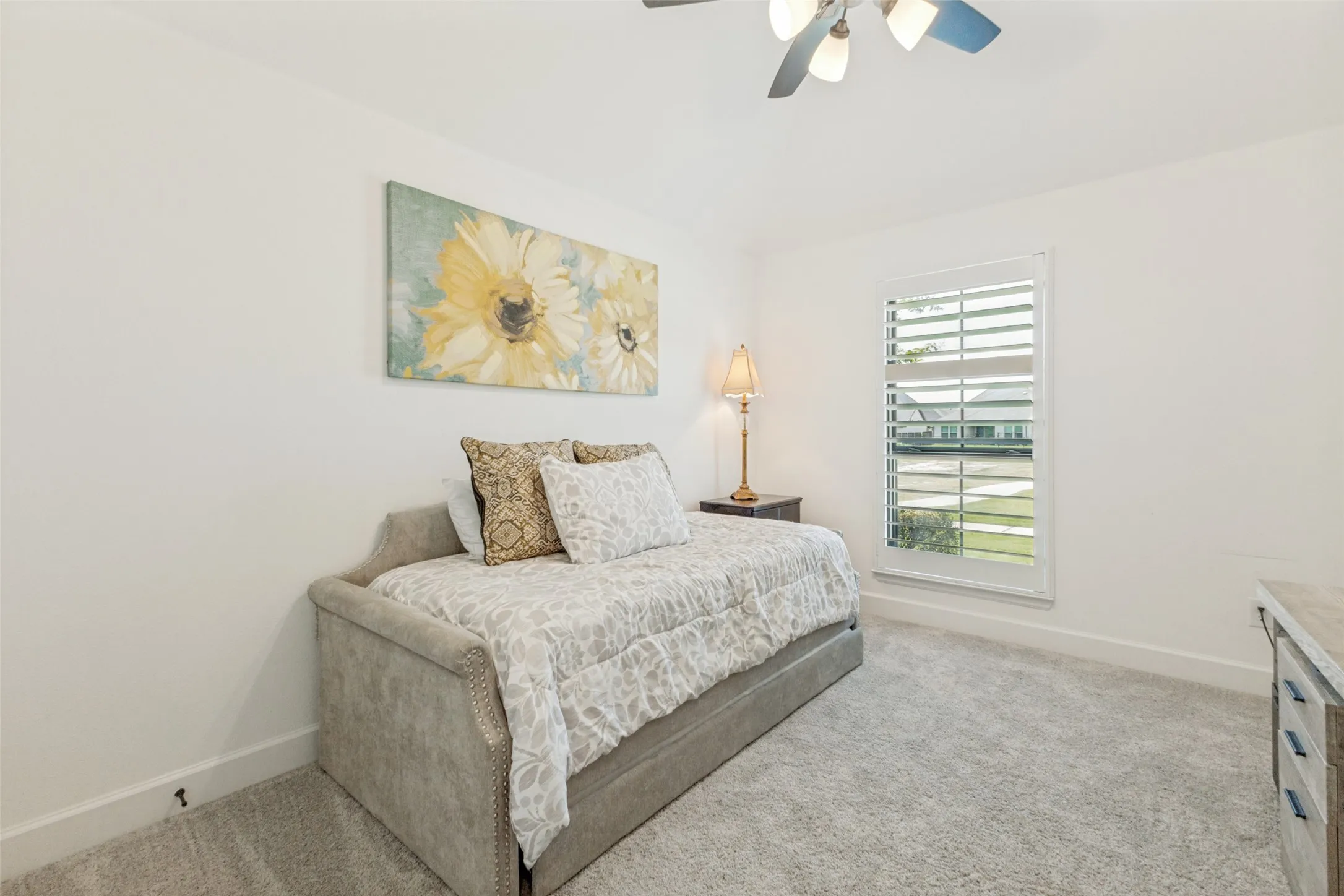 White walls, ceiling fan with integrated lighting, window with plantation shutters, light-toned carpeting, and a built-in dresser with a stone-finish top