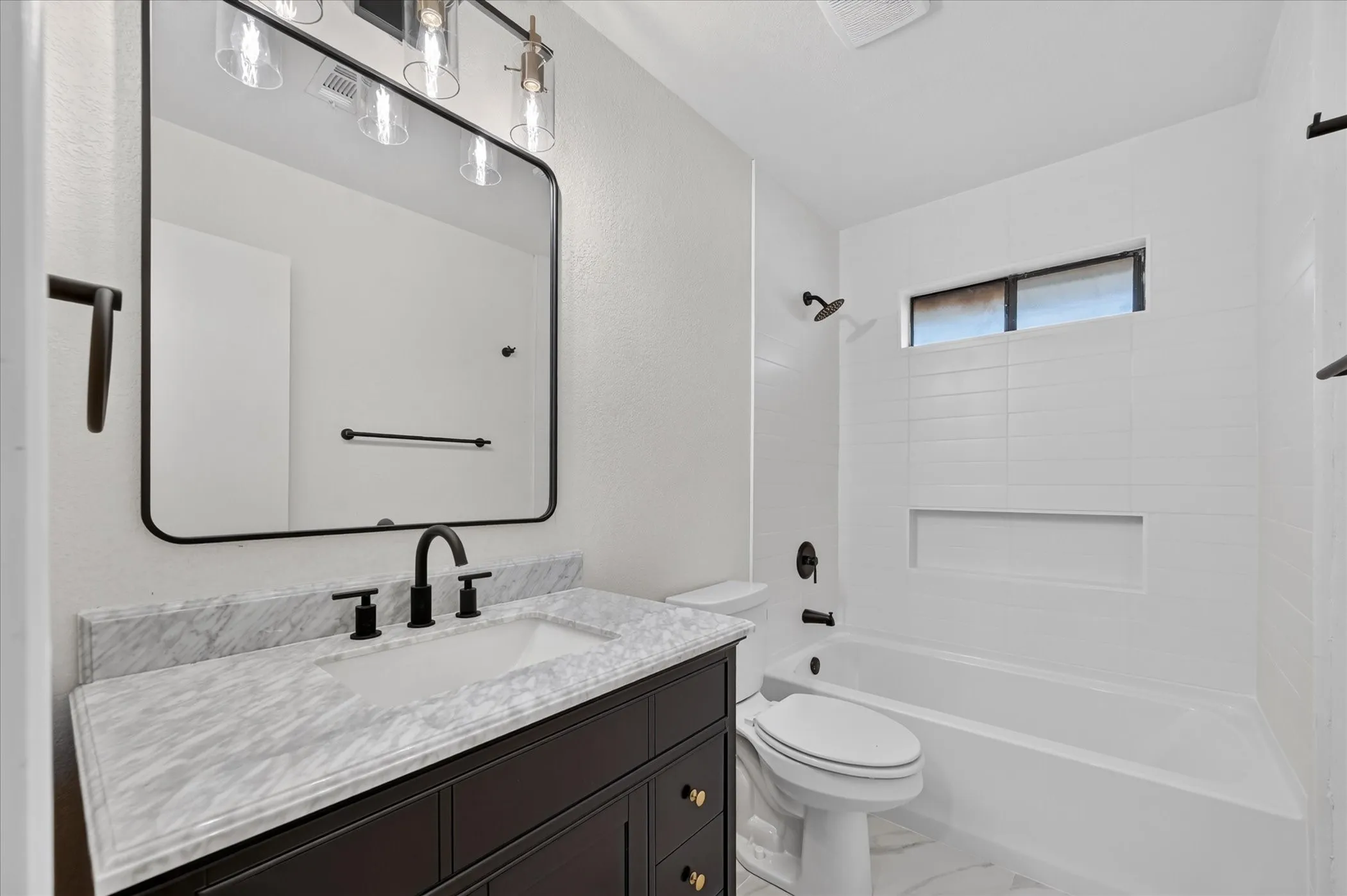 Rectangular vanity mirror with integrated lighting, dark wood vanity with gold-tone hardware, undermount sink with matte black fixtures, white tiled shower surround with built-in niche, and a window above the shower