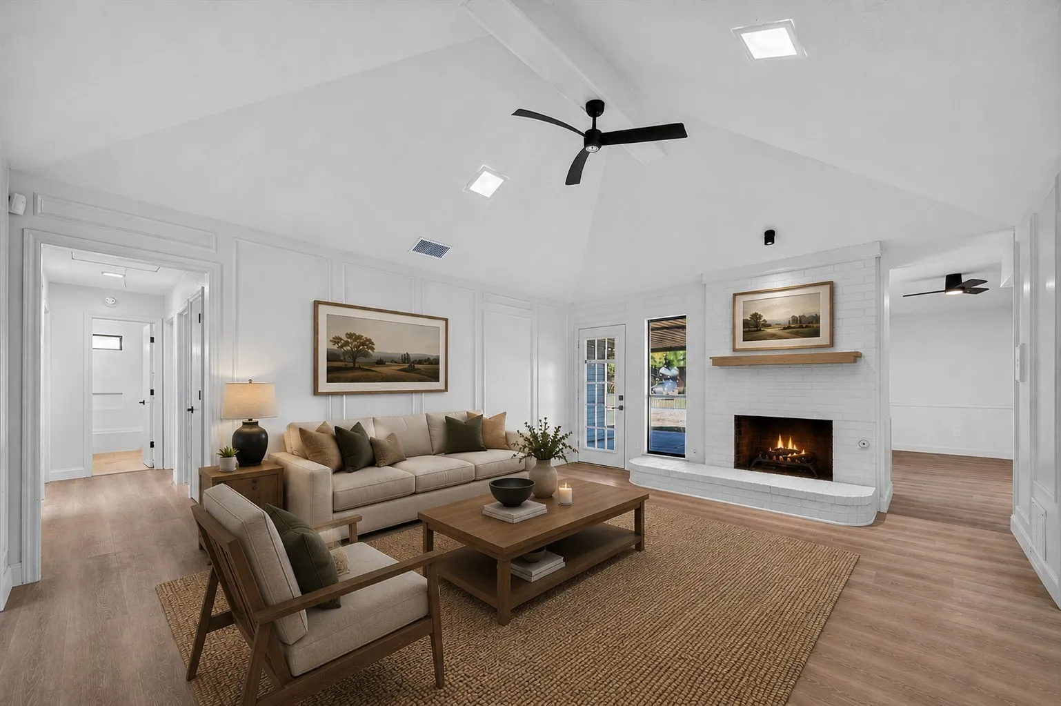 Vaulted white ceiling with exposed beam, white painted brick fireplace, wood-finish flooring, white paneling, and a ceiling fan