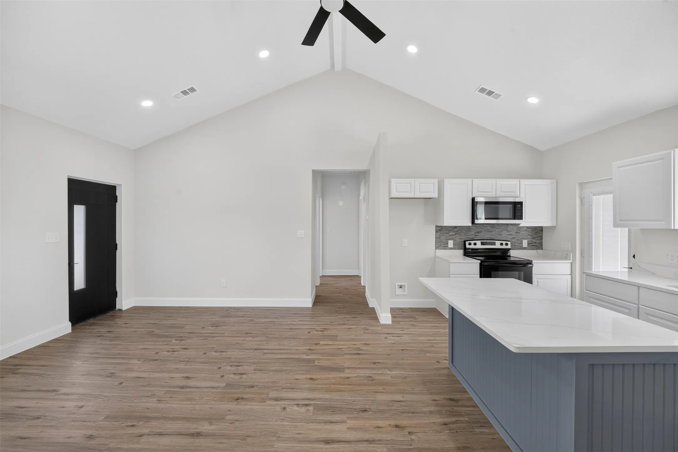 Vaulted ceiling with recessed lighting, wood-finish flooring, black front entry door, kitchen island with a light blue base, and white cabinetry
