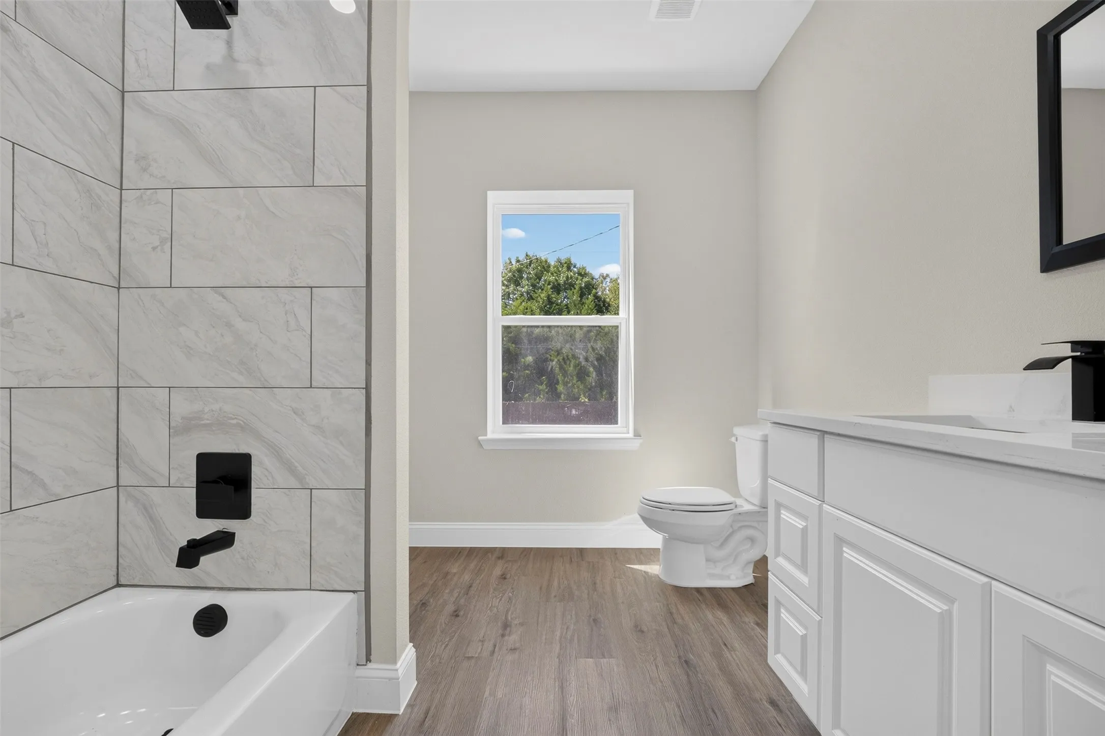 Bathroom featuring a bathtub with subway tile surround, matte black fixtures, wood-finish flooring, a single window, and a vanity with white cabinetry