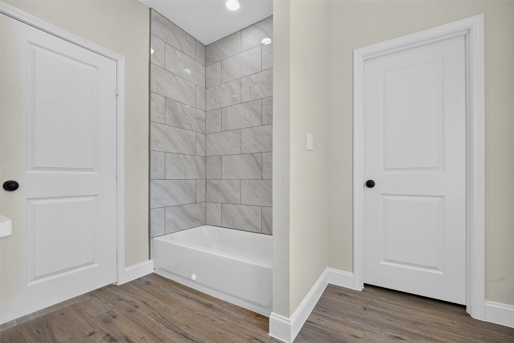 Primary bathroom featuring a soaking tub with tile surround, walk-in shower enclosure with marble-look tile walls, wood-finish flooring, recessed lighting, and dual white six-panel doors providing entry and access to adjoining spaces