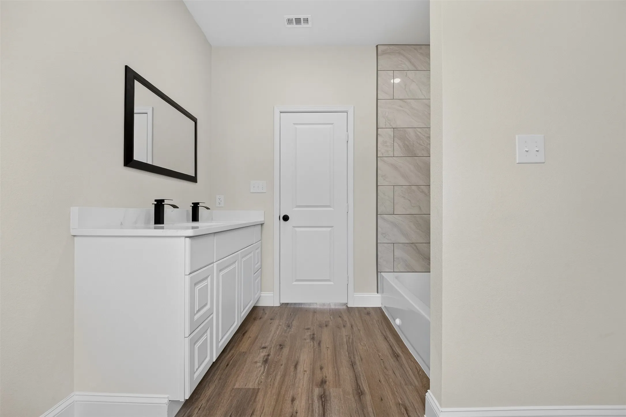 Dual sink vanity with white cabinetry, wood-finish flooring, black framed mirror, and tiled bath surround