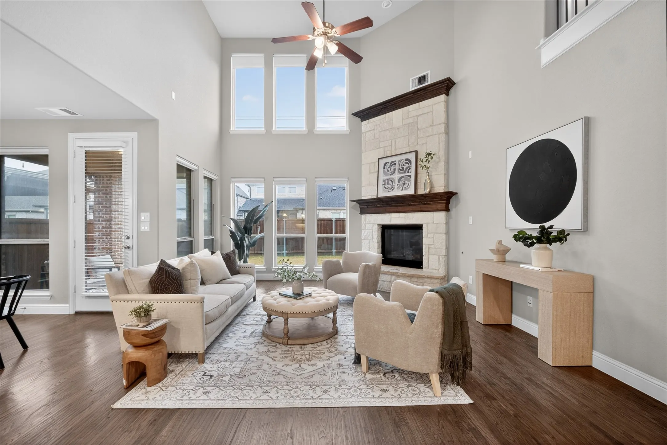 Two-story living area with high ceilings, featuring a stone fireplace with a substantial wood mantle, multiple tall windows, a ceiling fan, and wood-finish flooring