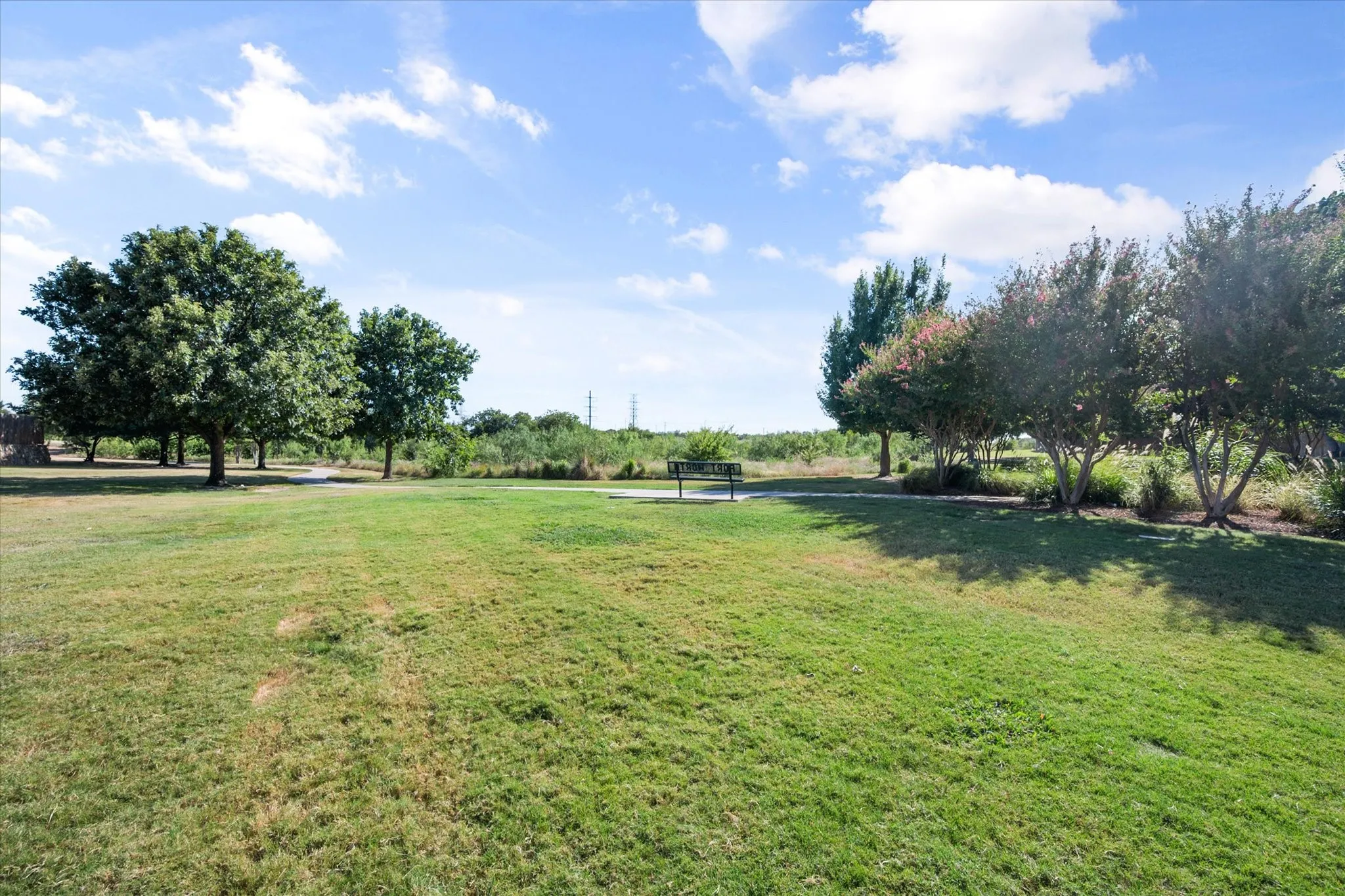 Expansive green space featuring mature trees, a paved pathway, and a park bench