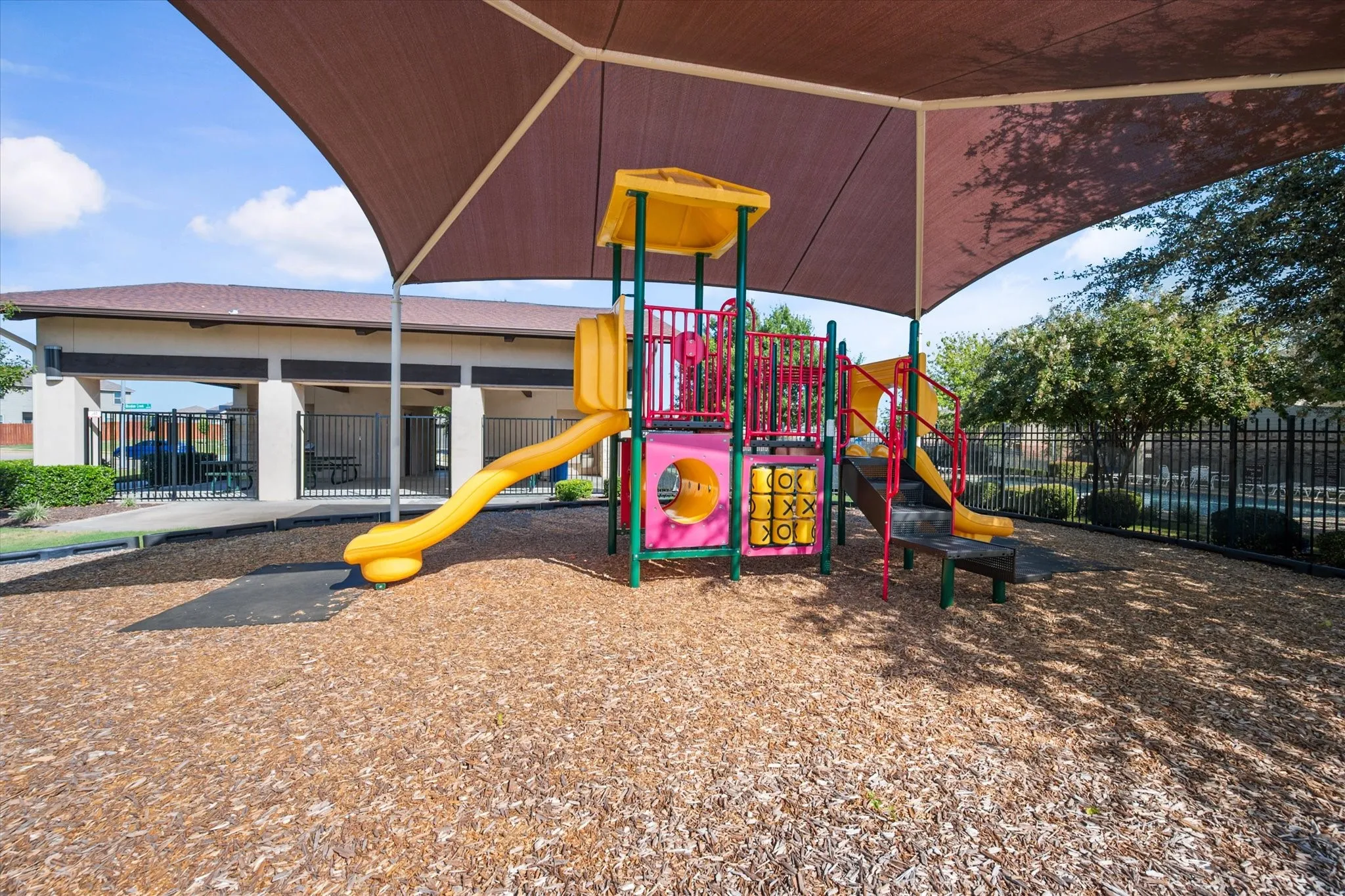 Playground structure with slides and climbing elements, covered by a large shade canopy, set on wood chip surfacing